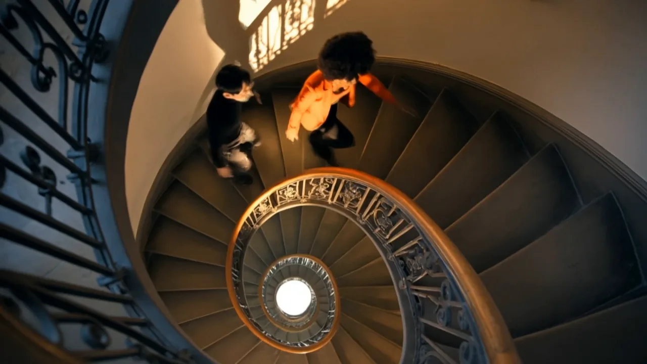 Vue plongeante dans un escalier en colimaçon : deux personnages, dont une femme en veste orange, dévalent les marches autour d'une rampe en fer forgé.